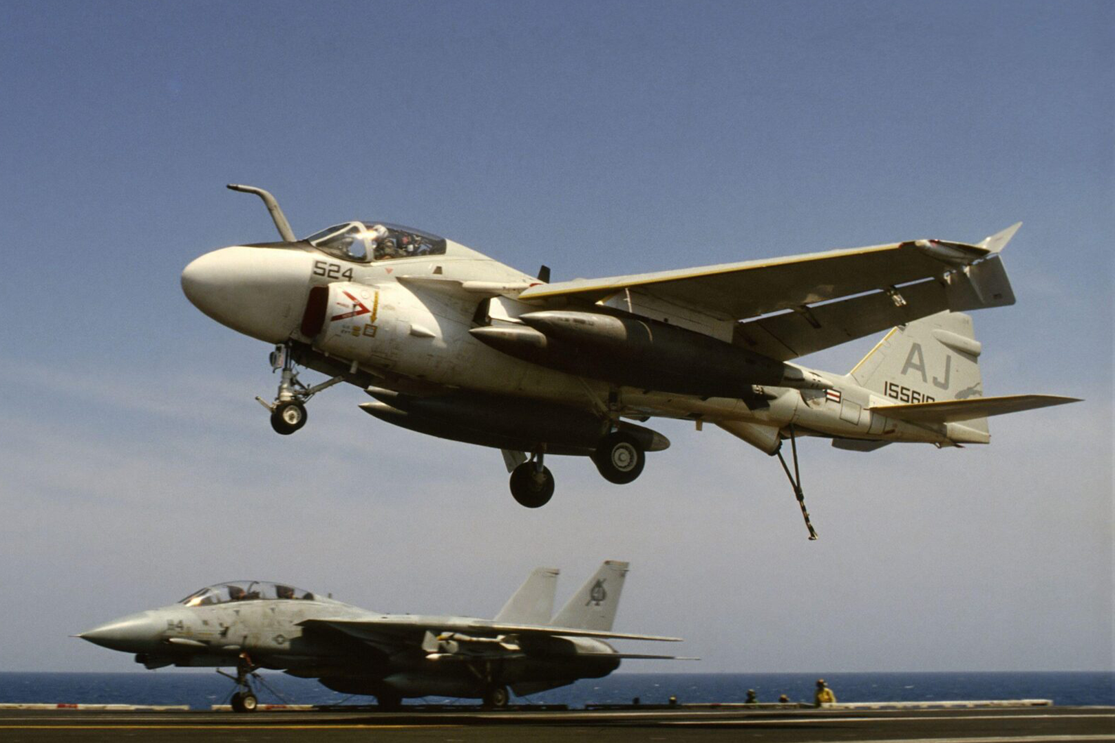 A jet plane flying over another jet on an aircraft carrier.